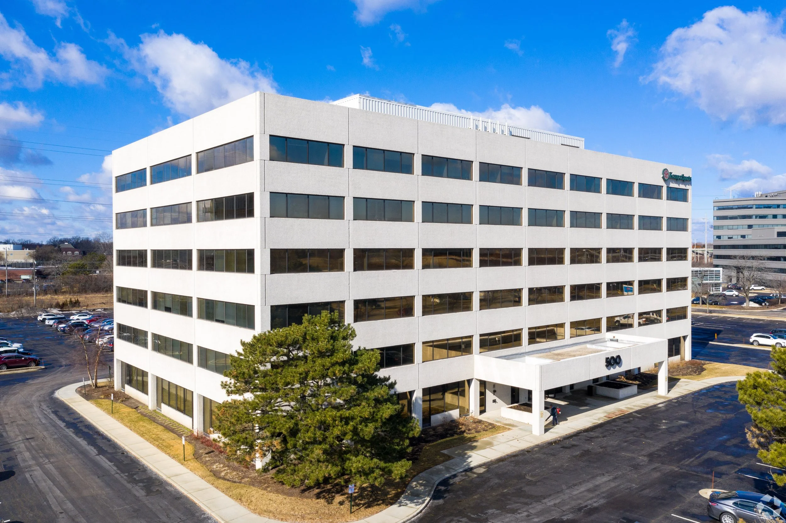 A multi-story office building with a white exterior and large rectangular windows, labeled with the number 500, situated in a parking lot with several cars and some trees under a blue sky with clouds.