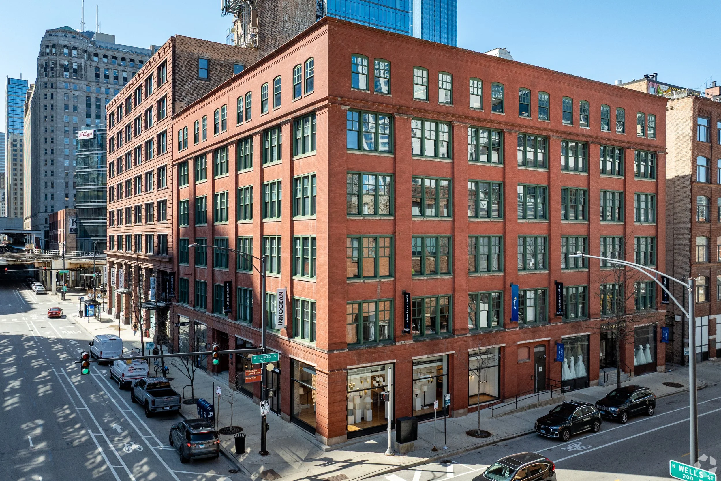 A multi-story red brick building on a city street corner with retail stores on the ground floor and large windows on upper floors. Vehicles are parked along the street and traffic lights are visible at the intersection.