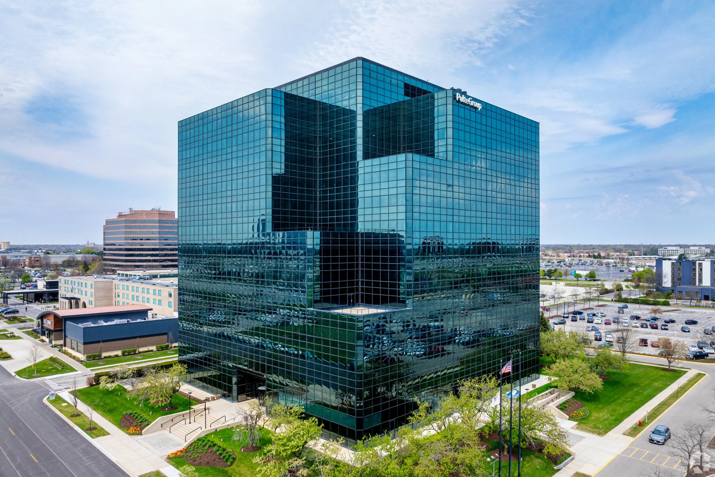 A modern glass office building with blue-tinted windows surrounded by parking lots and greenery under a partly cloudy sky.