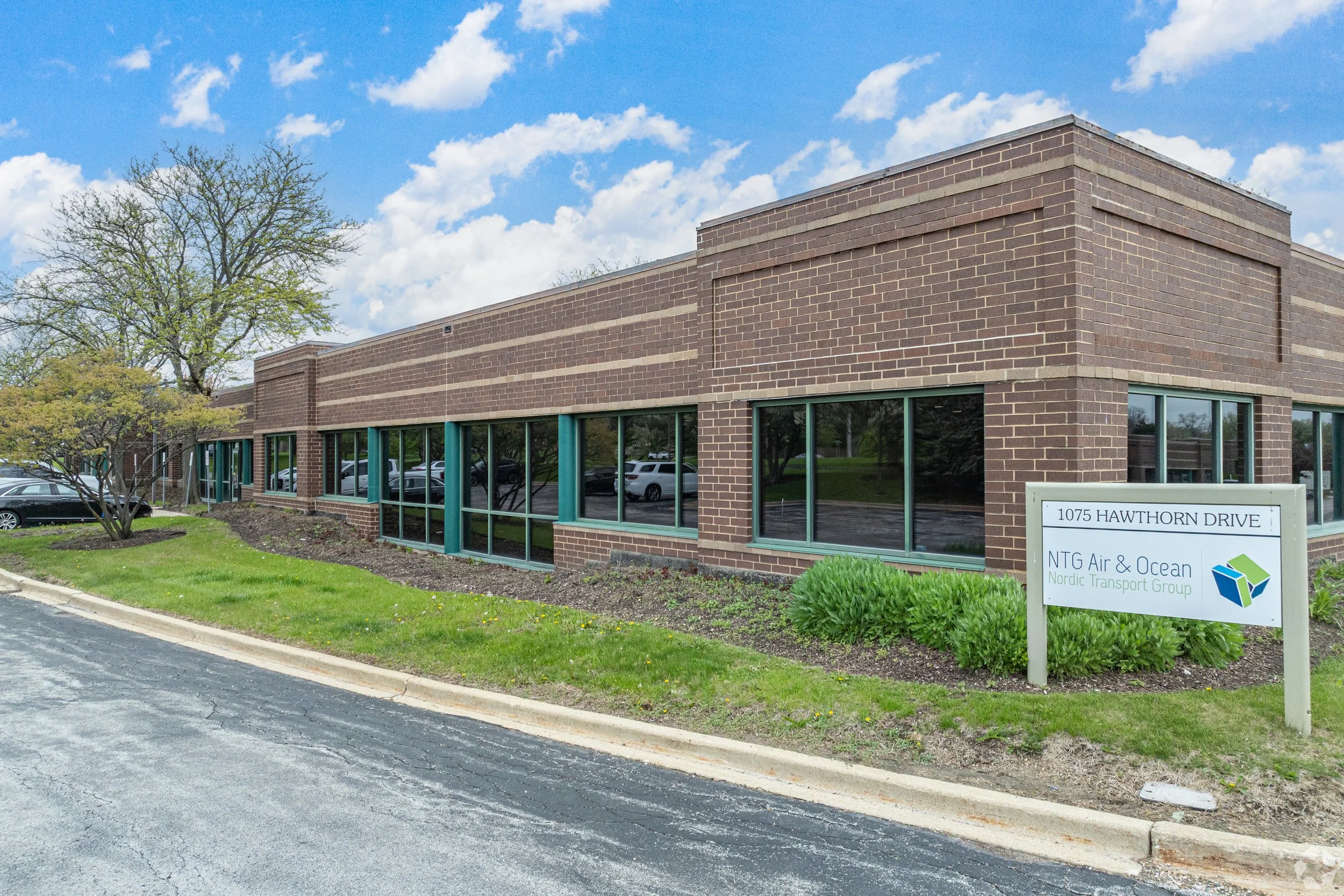 A brick building with large windows reflecting parked cars and trees. A sign in front reads "1075 Hawthorn Drive, NTG Air & Ocean, Nordic Transport Group."
