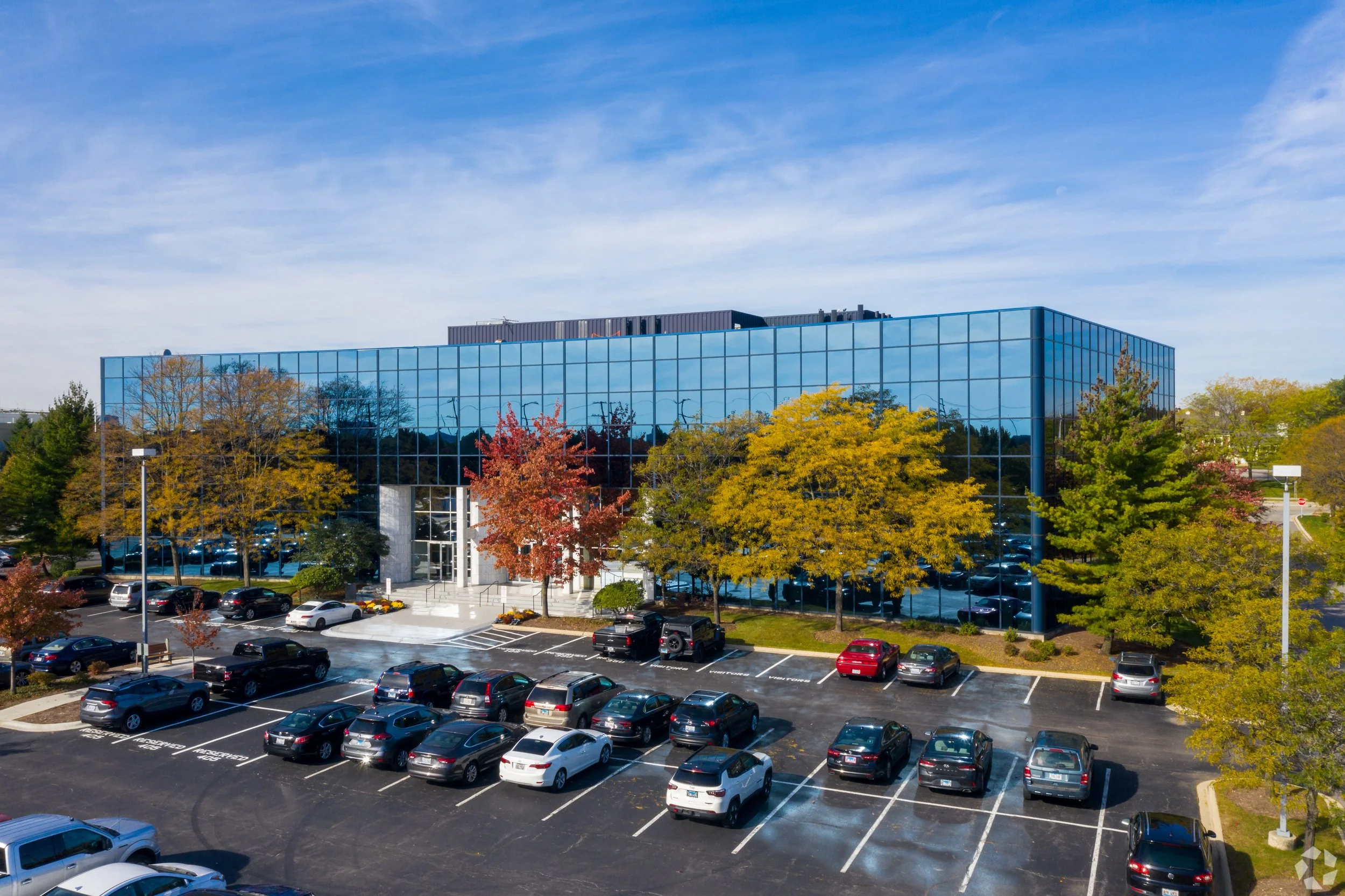 Modern glass office building surrounded by trees with colorful autumn leaves, parking lot filled with cars, under a partly cloudy blue sky.