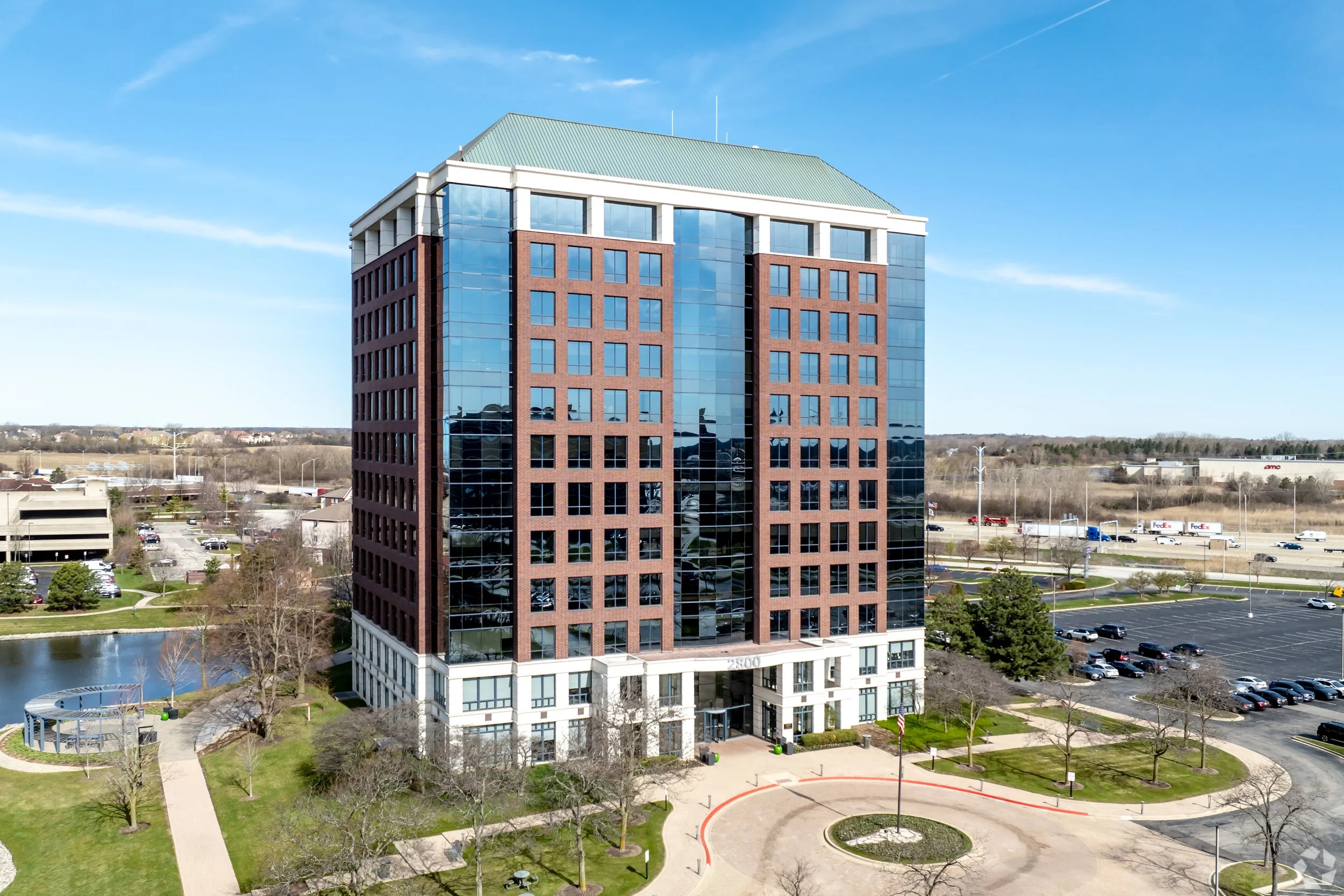 A tall office building with a modern glass and brick exterior, surrounded by a park with walking paths, trees, and a pond, under a clear blue sky.
