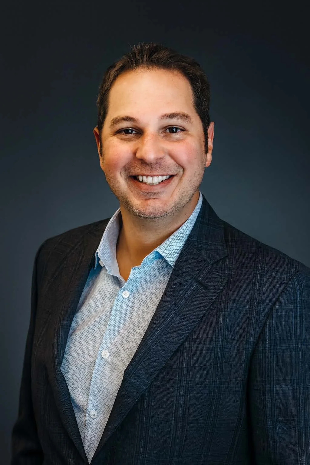 Professional headshot of a smiling man wearing a dark plaid blazer and a light blue collared shirt against a dark background.