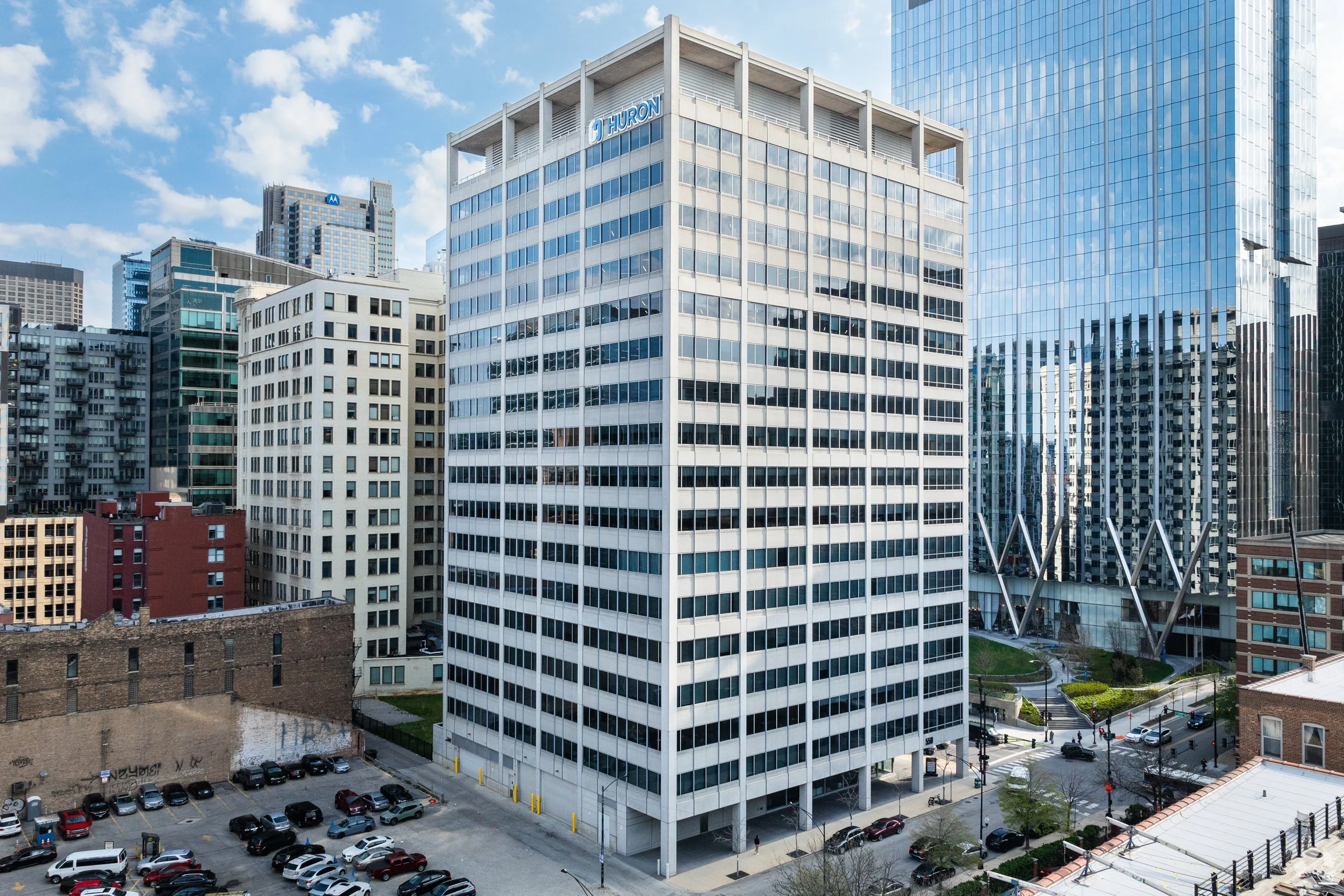 Tall white office building with the Huron logo on top, surrounded by other high-rise buildings in a cityscape, with parking lot in foreground and a blue sky with scattered clouds.