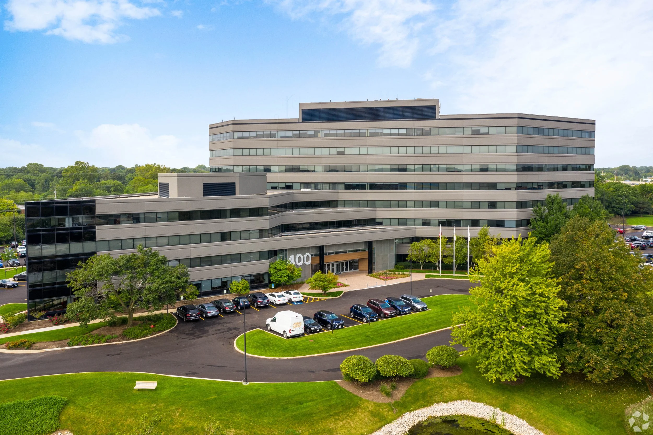 A modern, multi-story office building with a parking lot and surrounding greenery on a clear day.