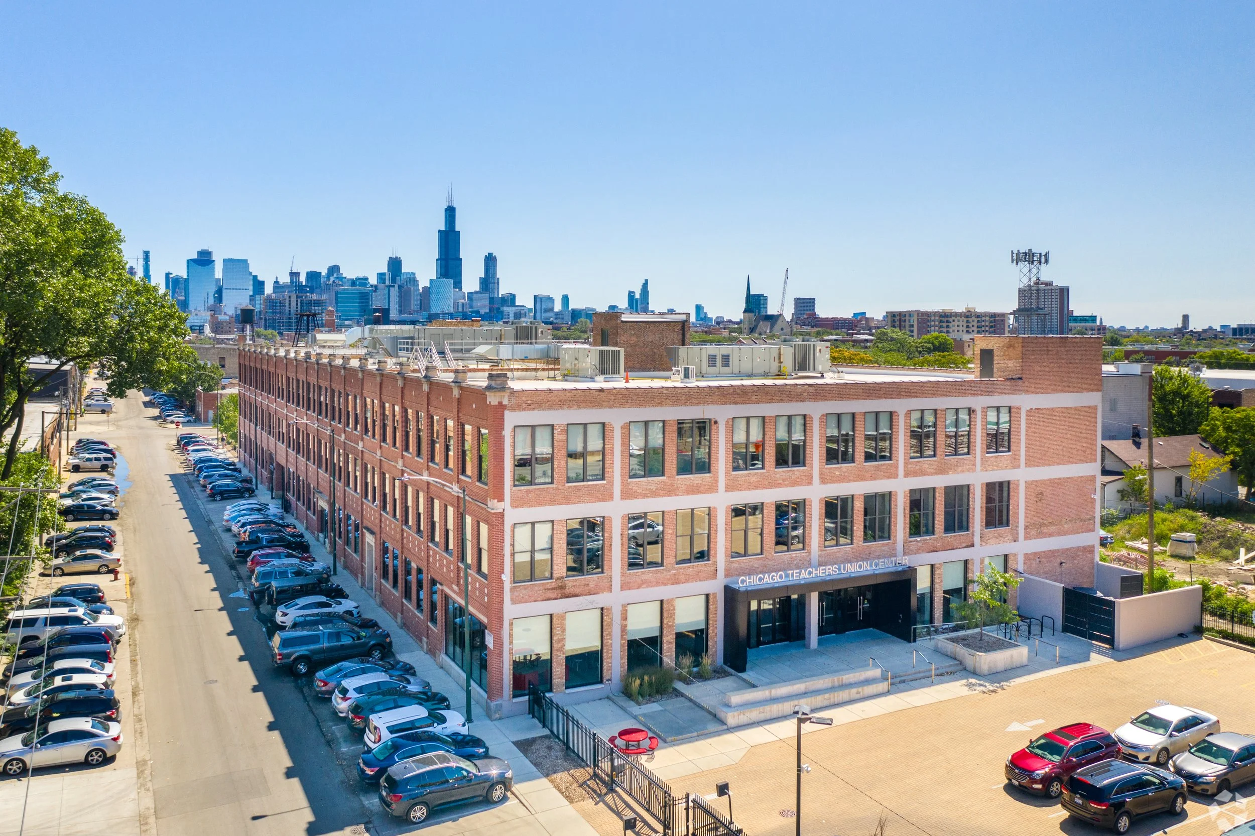A red brick building labeled Chicago Teachers Union Center with a parking lot in front, and Chicago skyline with Willis Tower in the background under a clear blue sky.