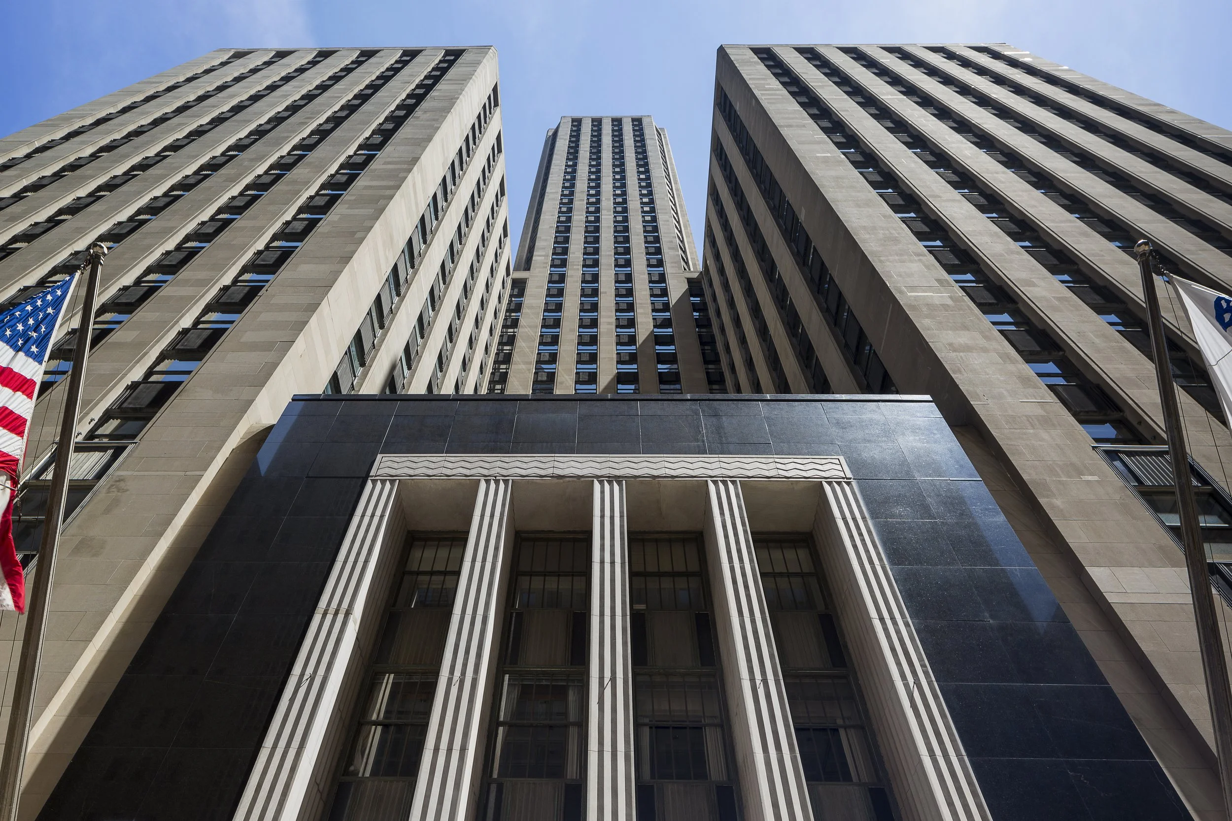 From a low angle view, tall skyscrapers with a mix of modern and classical architectural styles, American flags on poles in front, under a clear blue sky.