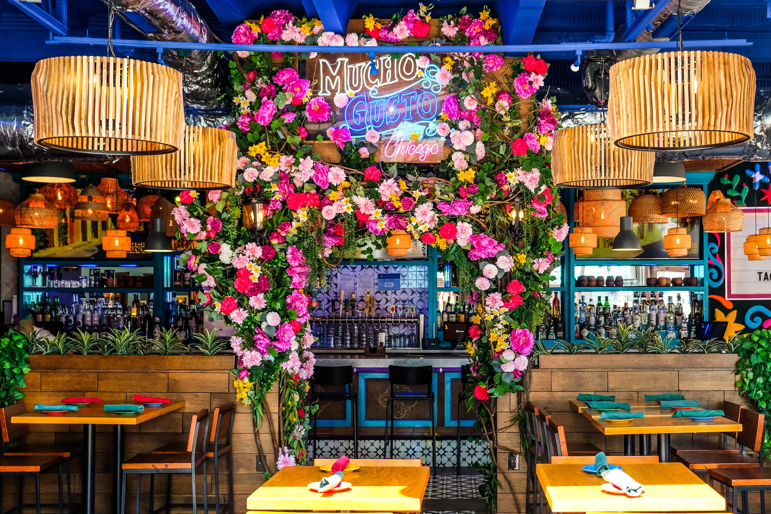 festive restaurant interiors with colorful flower arch, a neon sign, wooden light fixtures, and wooden tables and chairs