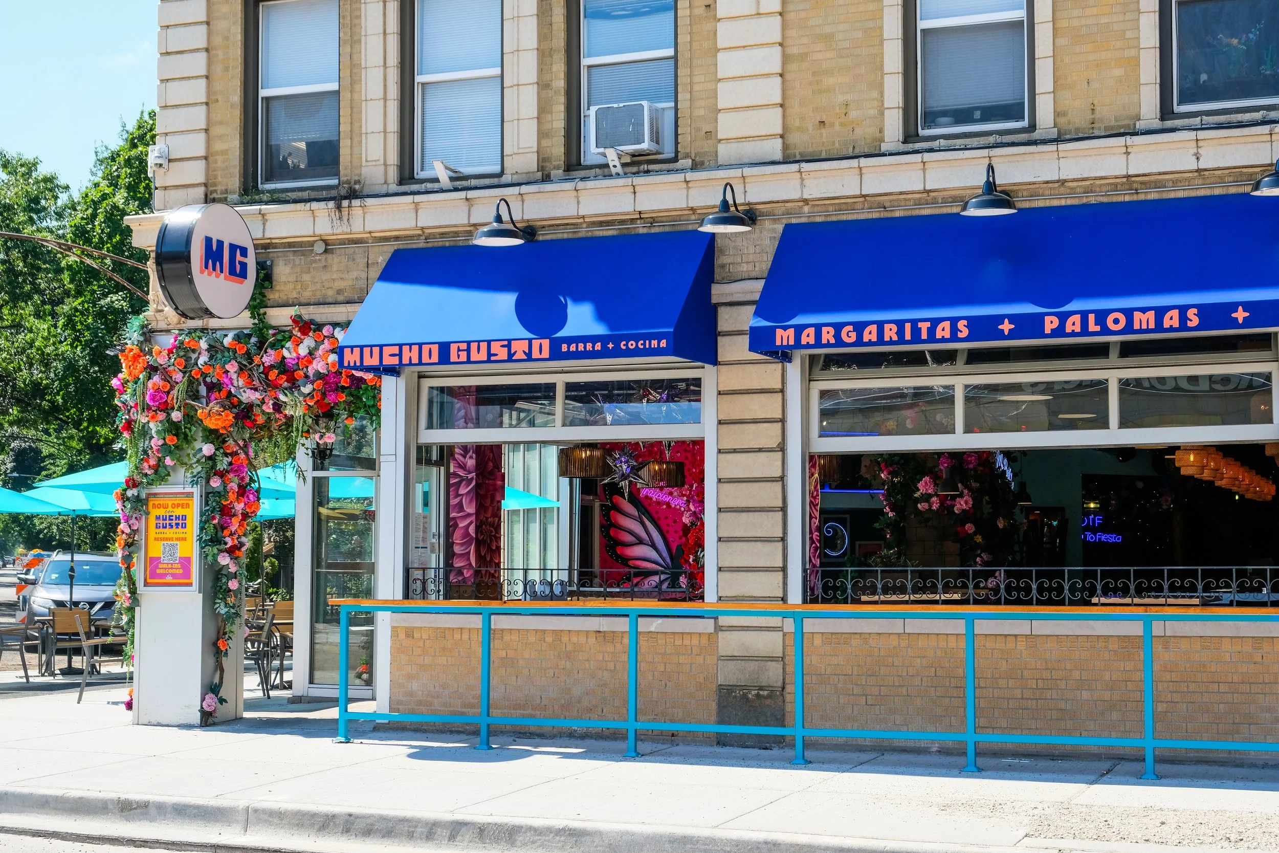 a restaurant's yellow brick exterior with blue awning with pink lettering reading Mucho Gusto Barra + Cocina, Margaritas and Palomas