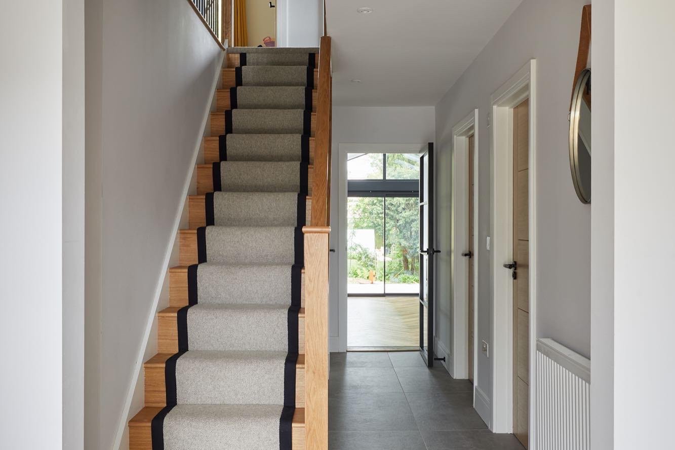 Interior view of a modern home hallway with stairs, doors, and a glass door leading outside, decorated with neutral tones and natural wood accents.