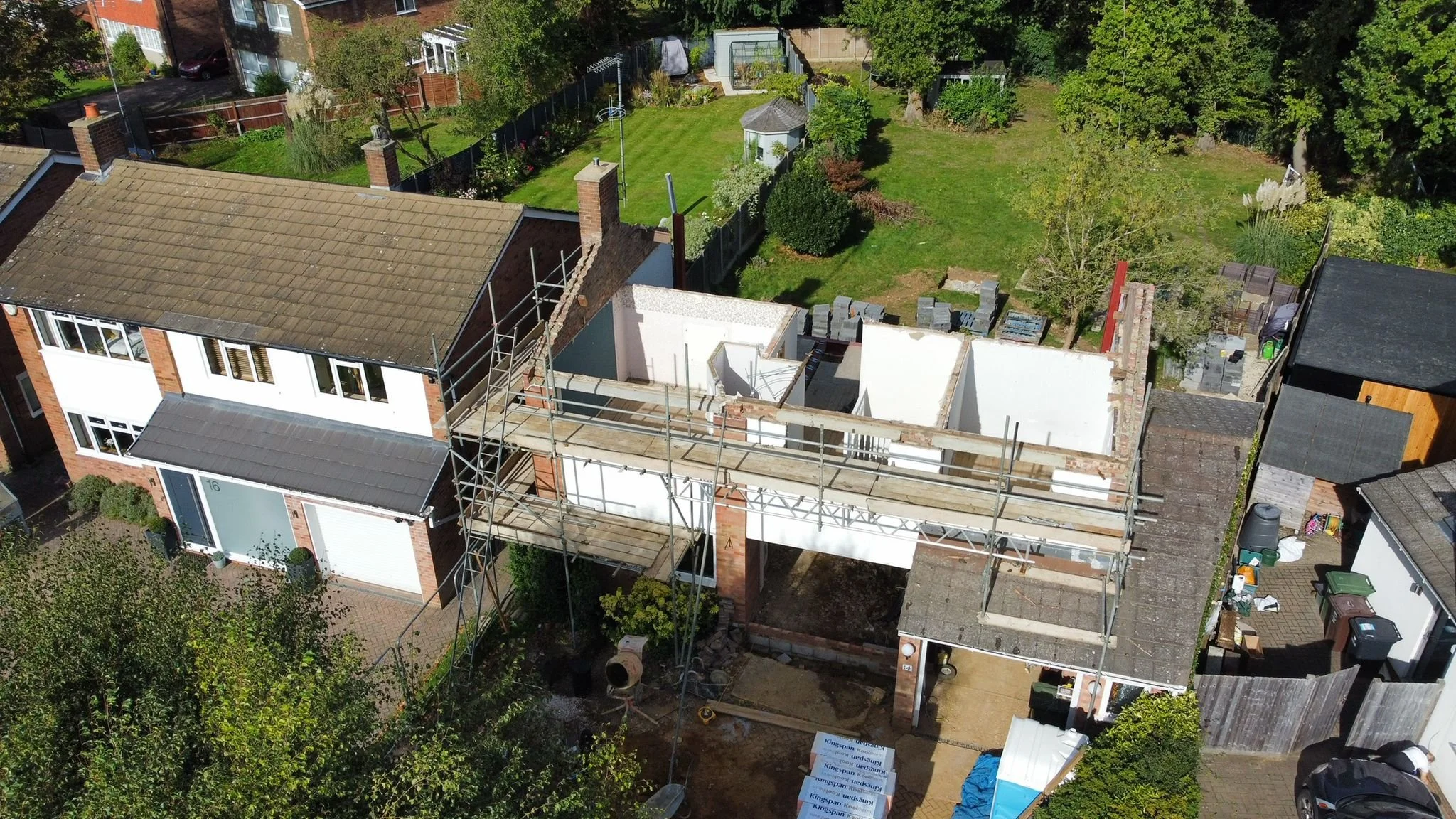 Aerial view of a two-story house under construction with scaffolding around it, neighboring houses, a backyard with a lawn, trees, and garden areas.