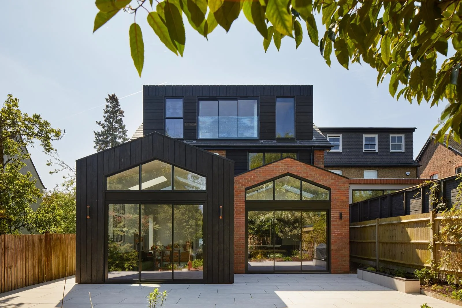 Modern multi-story house with black and red brick exterior, large glass windows, and a patio area surrounded by wooden fencing and trees.