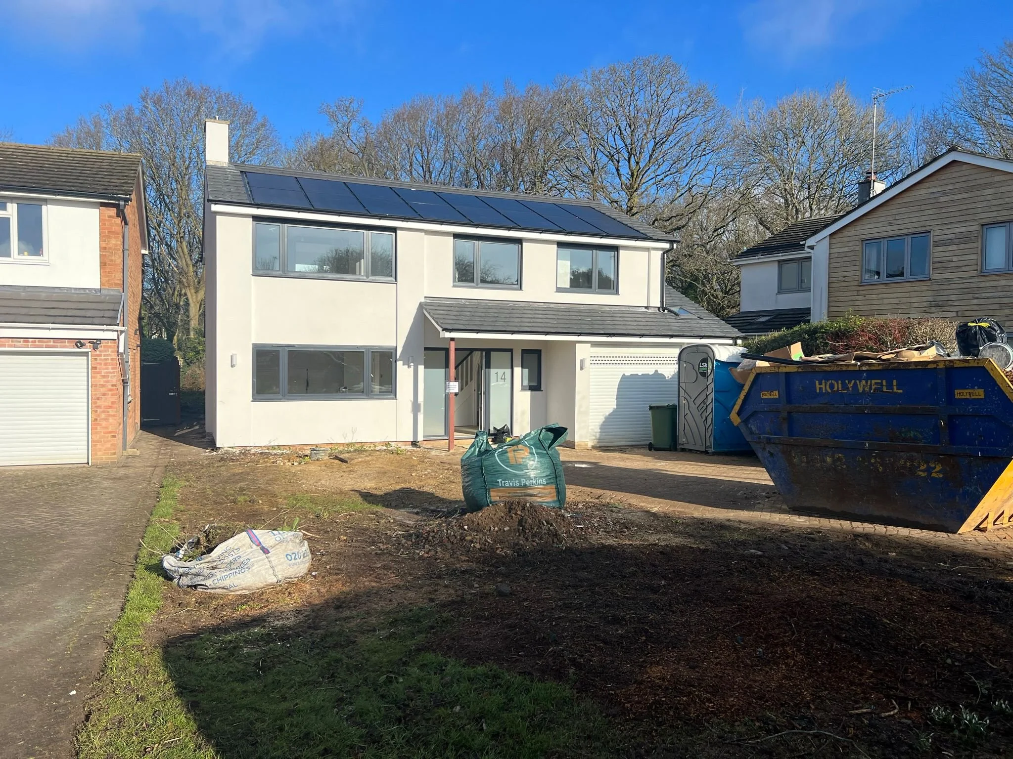 Construction site in front of a modern house with solar panels on the roof, surrounded by neighboring houses, piles of dirt, construction bags, and a large dumpster.