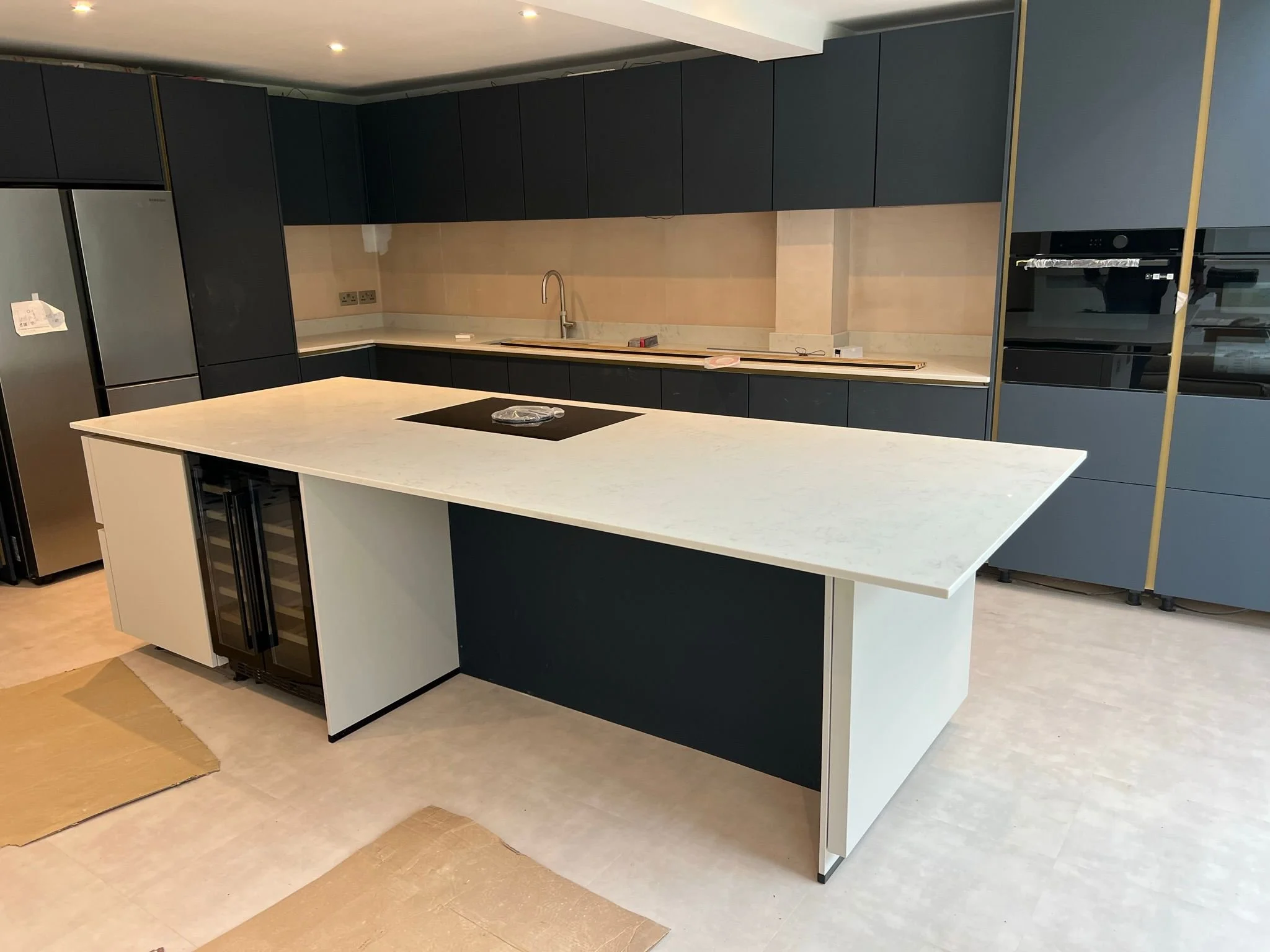 Modern kitchen with dark cabinets, a large white island with a cooktop, stainless steel appliances, and built-in ovens.