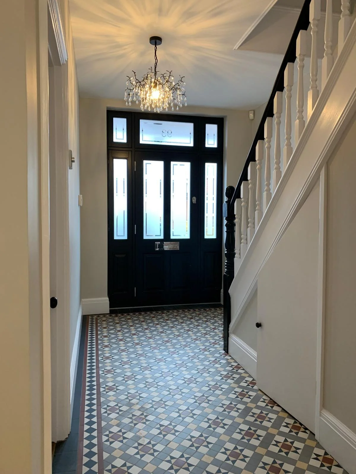 Entrance hallway with patterned tile floor, a black front door with frosted glass panels, a chandelier with crystal accents, and white stairs with black handrail.