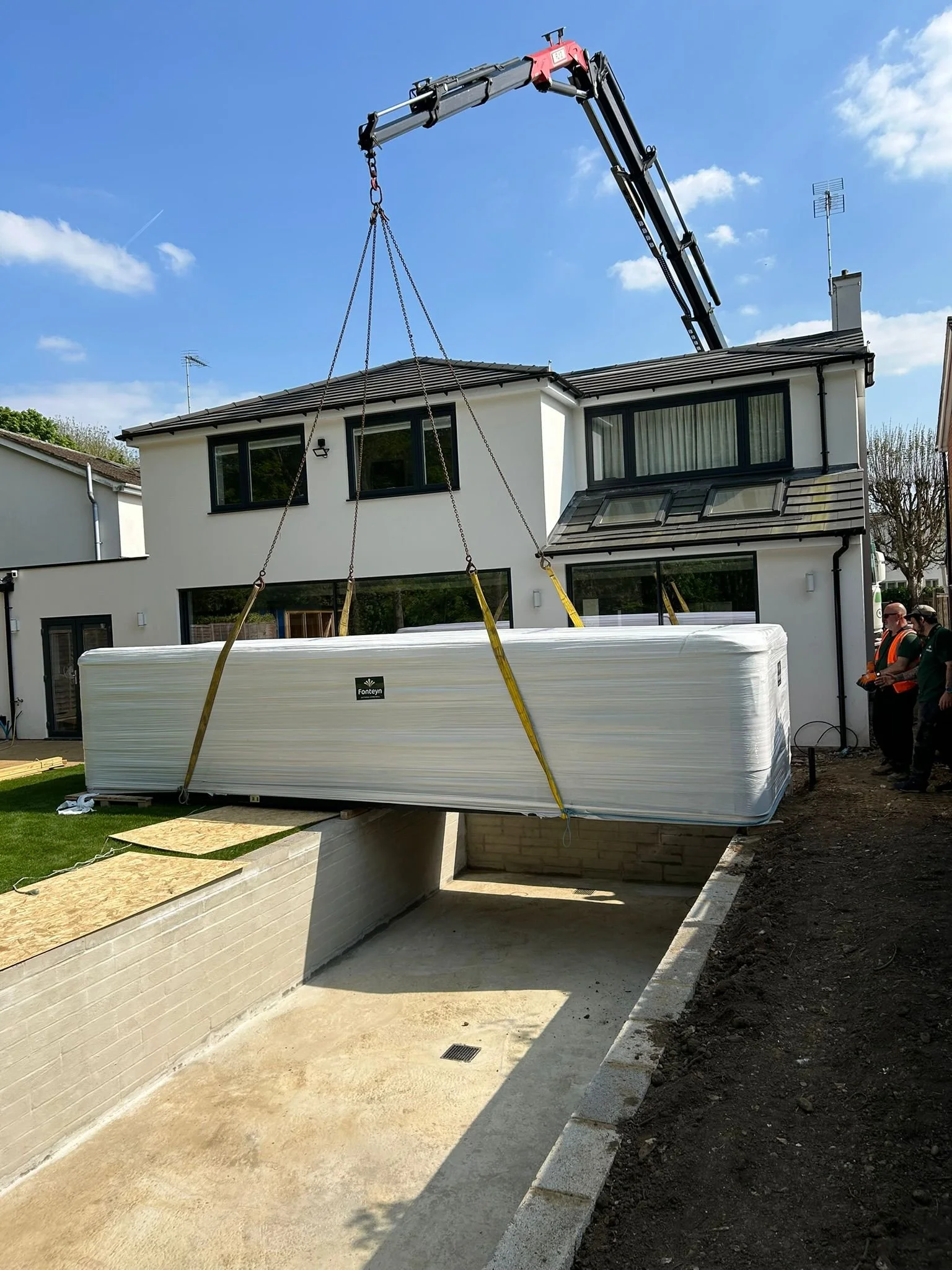 A crane lifting a large wrapped construction material into a basement of a modern two-story house with white exterior walls and black window frames during construction, with workers observing nearby.