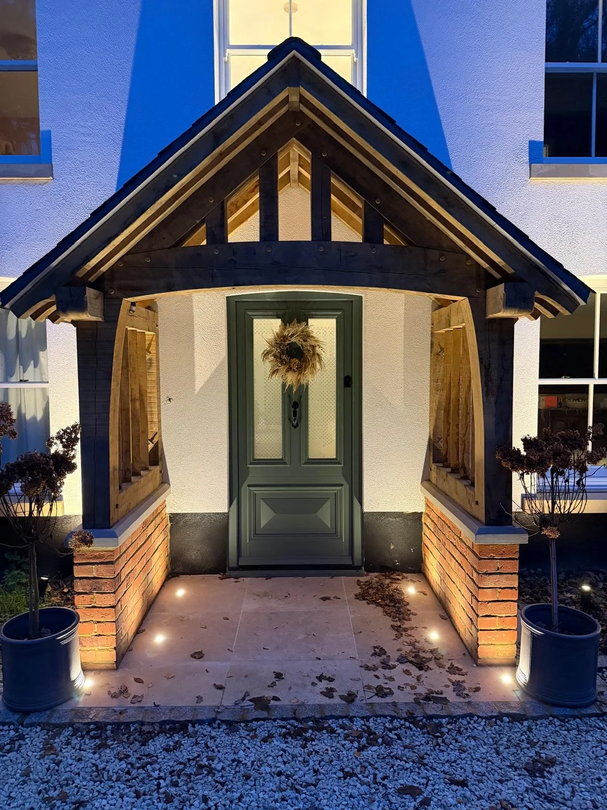 Front porch of a house with a green door decorated with a wreath, brick planters, potted trees, and pathway lighting.