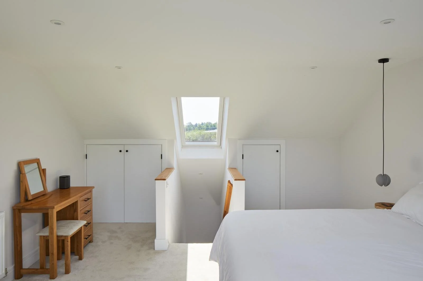 A bedroom with white walls, a bed with white bedding on the right, a wooden dresser with a mirror and a small speaker on the left, a small wooden stool, and a skylight window on the ceiling.