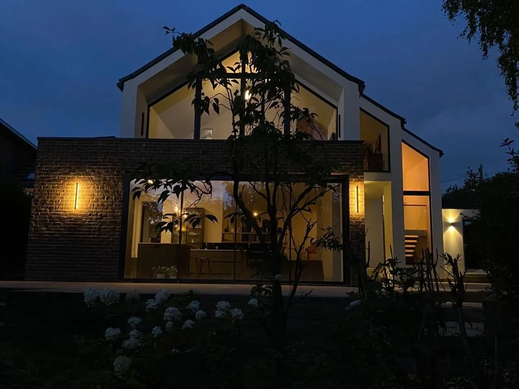 Modern two-story house illuminated at night, with large glass windows and brick accents, surrounded by trees and plants.