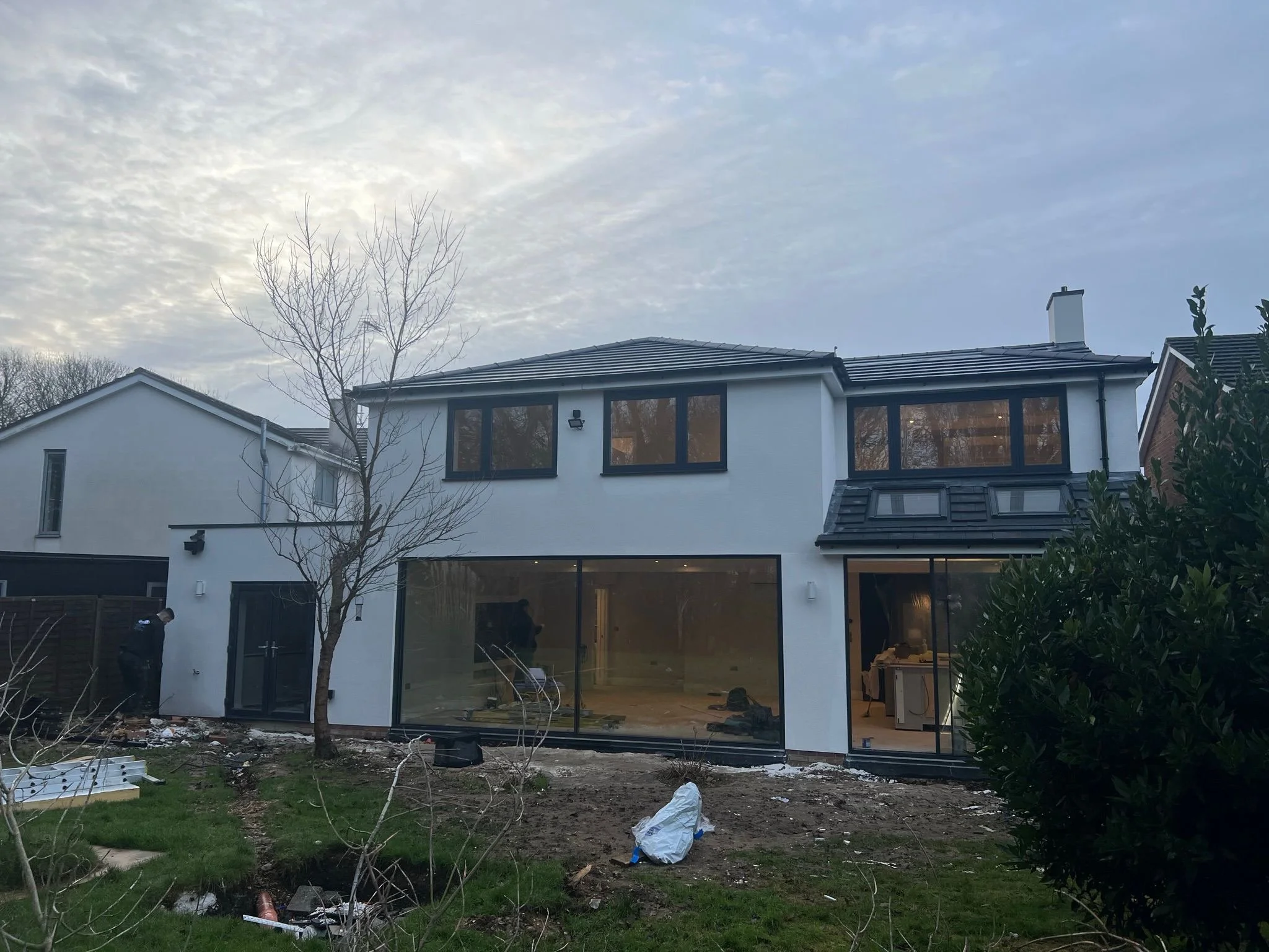 Newly constructed modern white house with large glass sliding doors at the back, multiple windows on upper level, and ongoing landscaping work in the yard.