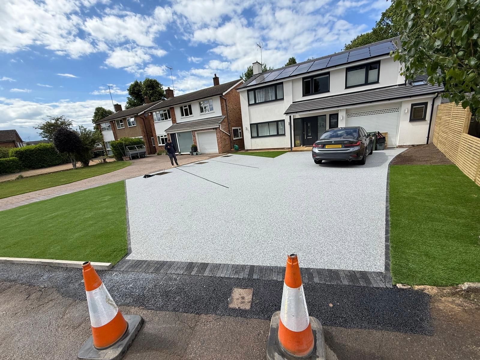 Residential driveway with newly laid light gray gravel, bordered by green grass and a brick pathway, with two orange traffic cones in the foreground, a black car parked near a white modern house, and other houses visible in the background under a partly cloudy sky.