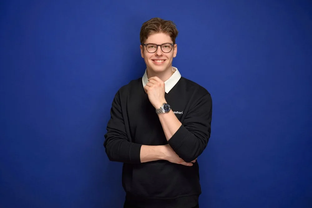 A young man with brown hair, glasses, and a watch, smiling and standing against a blue background, wearing a black sweater over a collared white shirt.