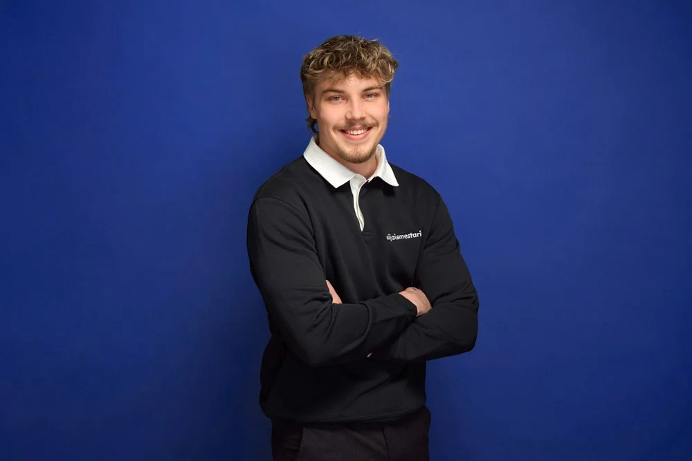 A young man with curly blond hair and a beard smiling, standing against a solid blue background, wearing a black long-sleeve shirt with a white collar.