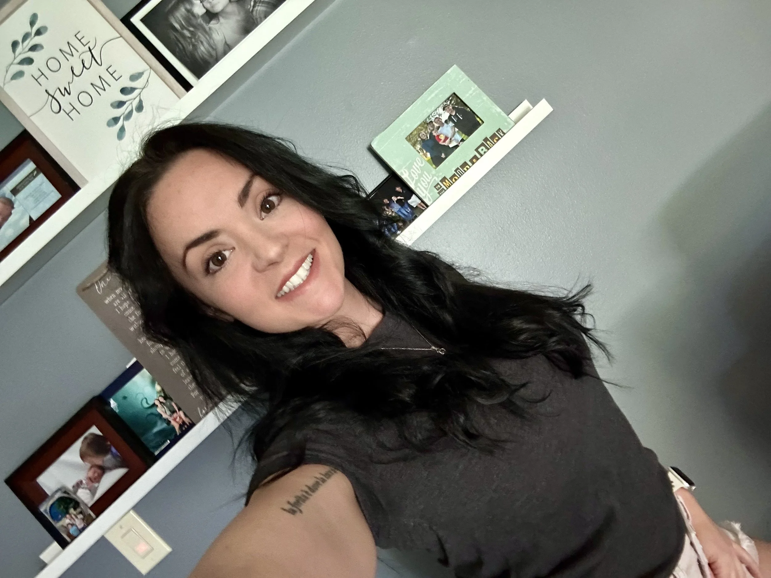 A woman with long black hair smiling for a selfie in front of a wall with framed photos and decorative signs.