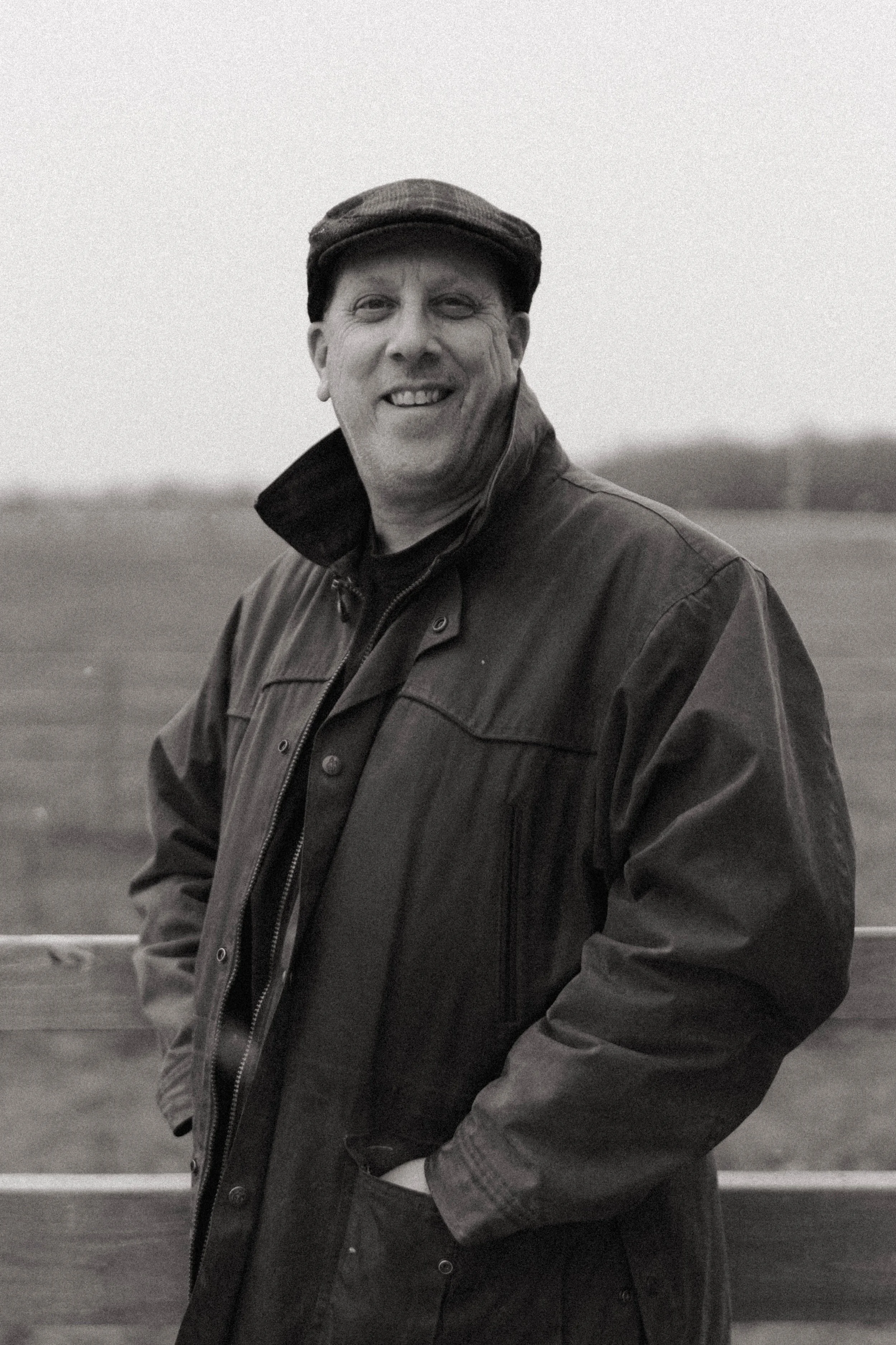A smiling man in a cap and jacket standing outdoors near a wooden fence with a blurred landscape in the background.