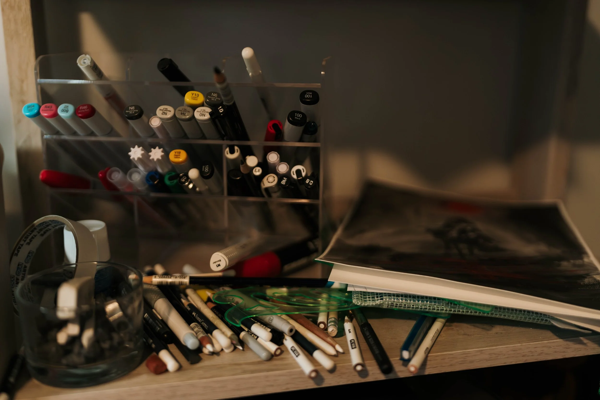 Art supplies including markers, pens, and paper scattered on a wooden surface with organized markers in a plastic container.