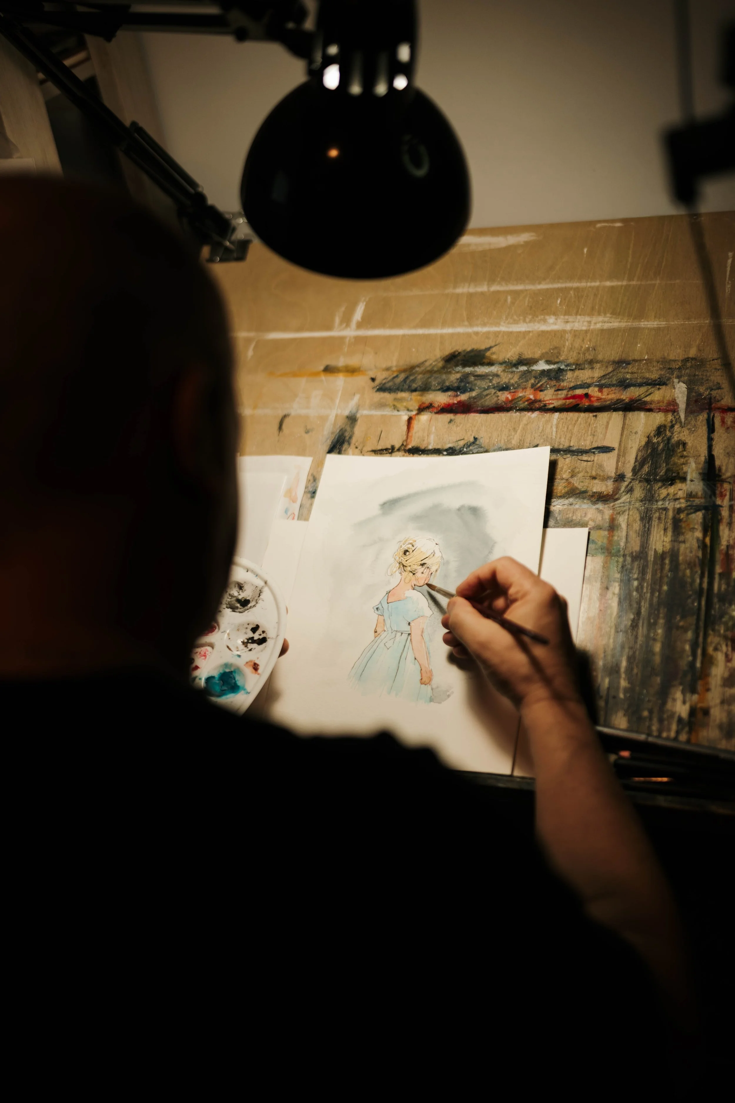 An artist painting a watercolor portrait of a young girl with blonde hair styled in an updo, wearing a blue dress, on a wooden desk with paint supplies.