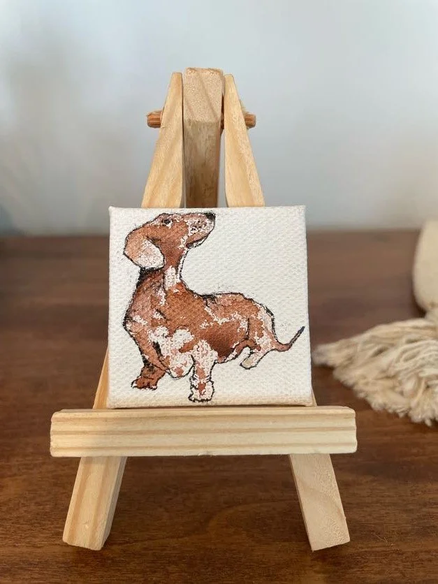 Miniature cross-stitch embroidered picture of a brown dog on a small wooden easel, placed on a wooden surface.