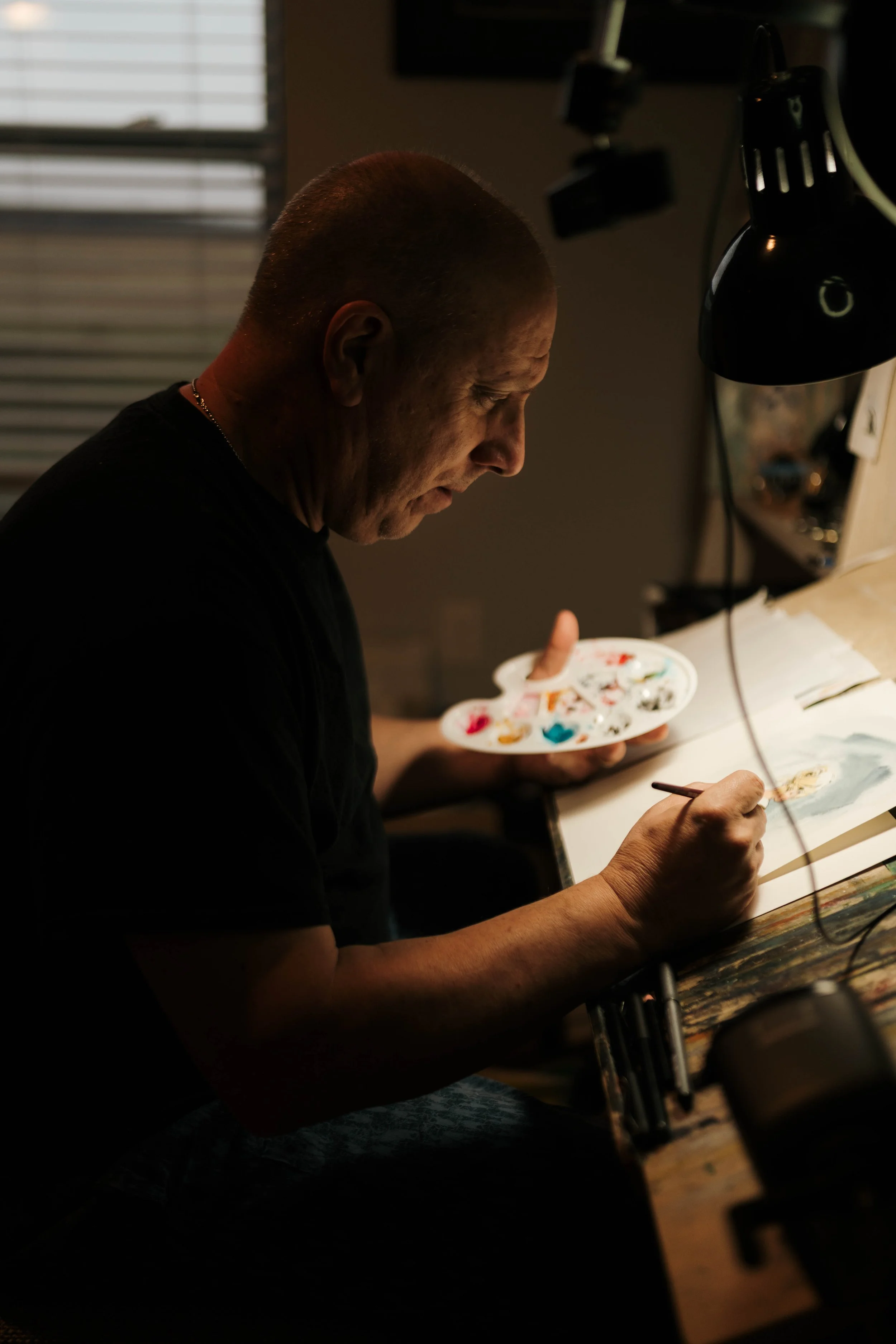 An artist painting at a desk under focused lighting in a dim room, holding a palette in one hand and a brush in the other.