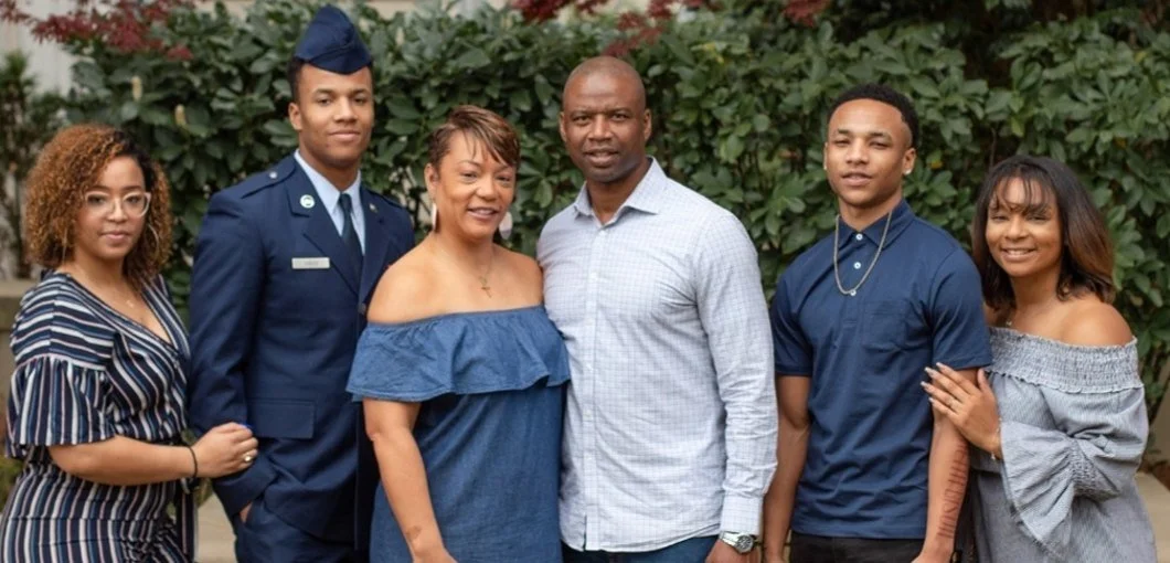 Group of six people standing outdoors in front of greenery, smiling at the camera, including a woman in a striped dress, a man in a military uniform, an older woman in a blue off-shoulder dress, a man in a light-colored shirt, a man in a dark-colored shirt with jewelry, and a woman in an off-shoulder top.