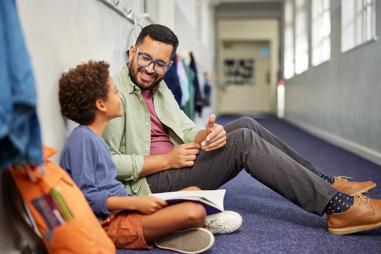 An adult man and a young boy sitting on the hallway floor, having a conversation and smiling at each other.