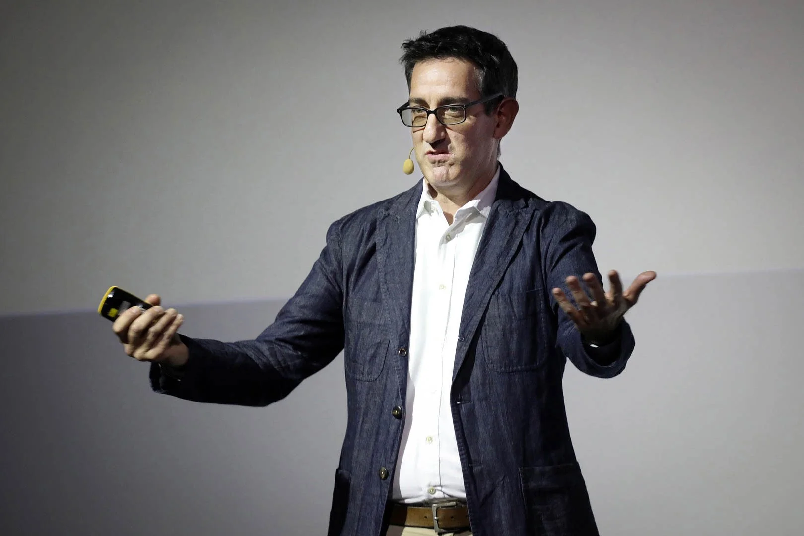Mark Maynard speaking with glasses and dark hair, wearing a white shirt and dark blazer, gesturing with one hand while holding a microphone headset and a yellow device in the other, standing against a plain background.