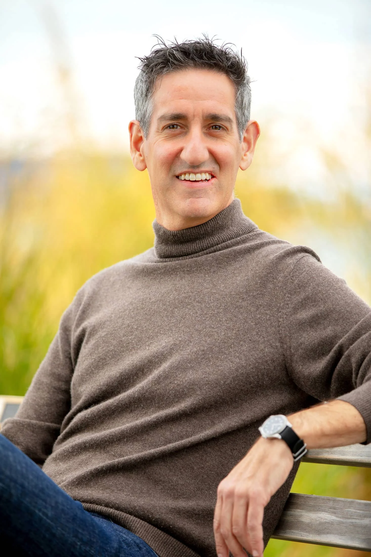 Mark Maynard sitting on a wooden bench outdoors with yellowing trees in the background, smiling at the camera.
