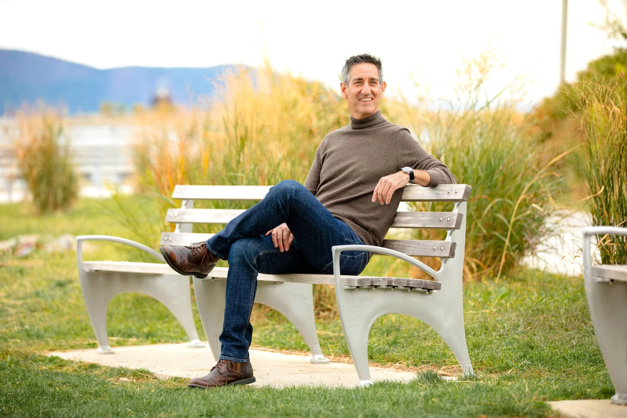 Mark Maynard sitting on a park bench in an outdoor setting during autumn, wearing a brown sweater, jeans, and brown boots, smiling and looking to the side.