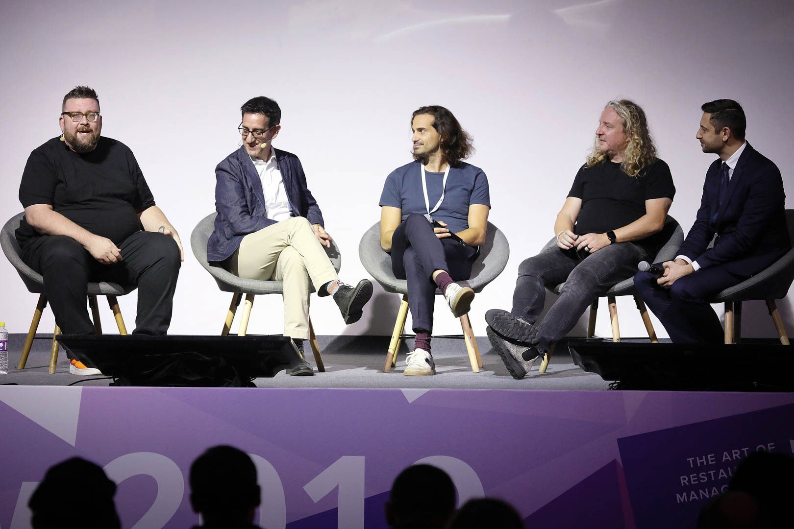Five men sitting on chairs on stage during a panel discussion, with a purple banner in front that reads 'The Art of Restaurant Management'. Mark Maynard as a featured guest on a panel