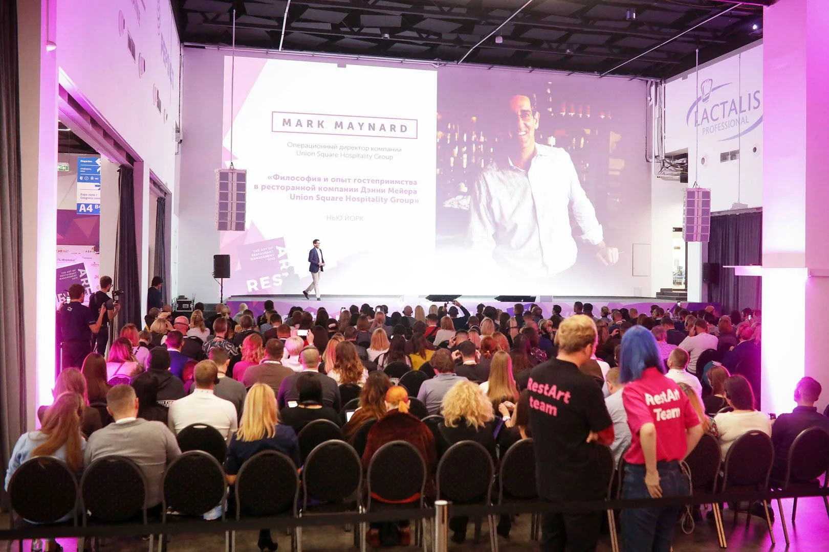 A large conference room filled with an audience watching a Mark Maynard presentation on stage. The stage has a large screen displaying text and a photo of a man in a white shirt.
