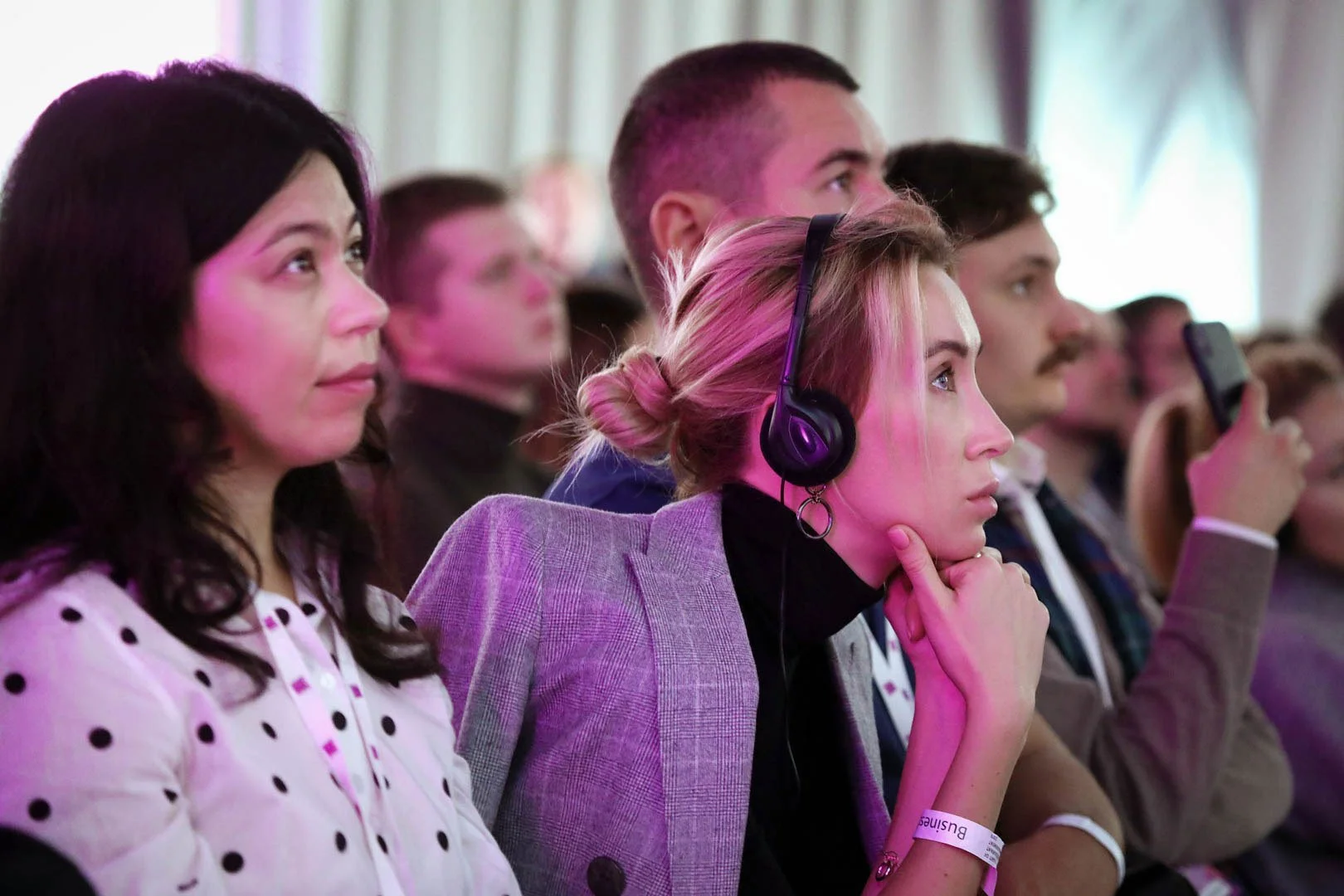 Group of people attending a conference or seminar, seated in rows, some taking notes or listening attentively, with a woman wearing headphones in the foreground.