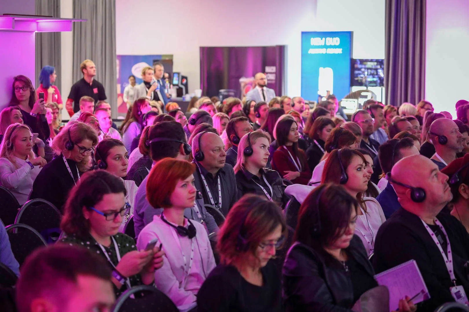 Crowd of people attending a conference, many wearing headsets, sitting in rows facing forward, with some individuals using smartphones, in a brightly-lit room with purple lighting and digital screens in the background.