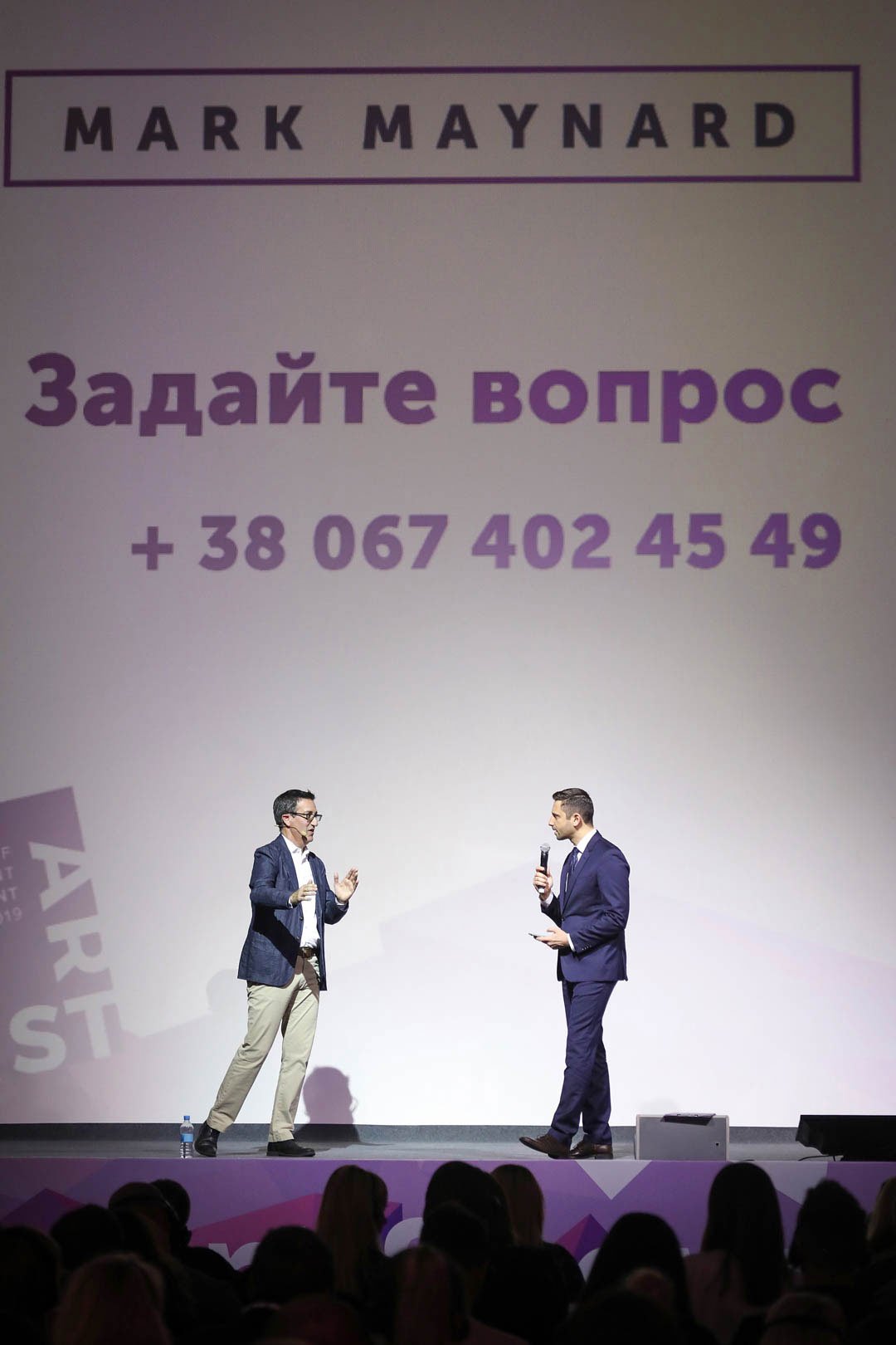 Mark Maynard on stage engaged in a conversation, with a large screen behind them displaying text in Ukrainian and a phone number. The audience is visible in silhouette at the bottom.