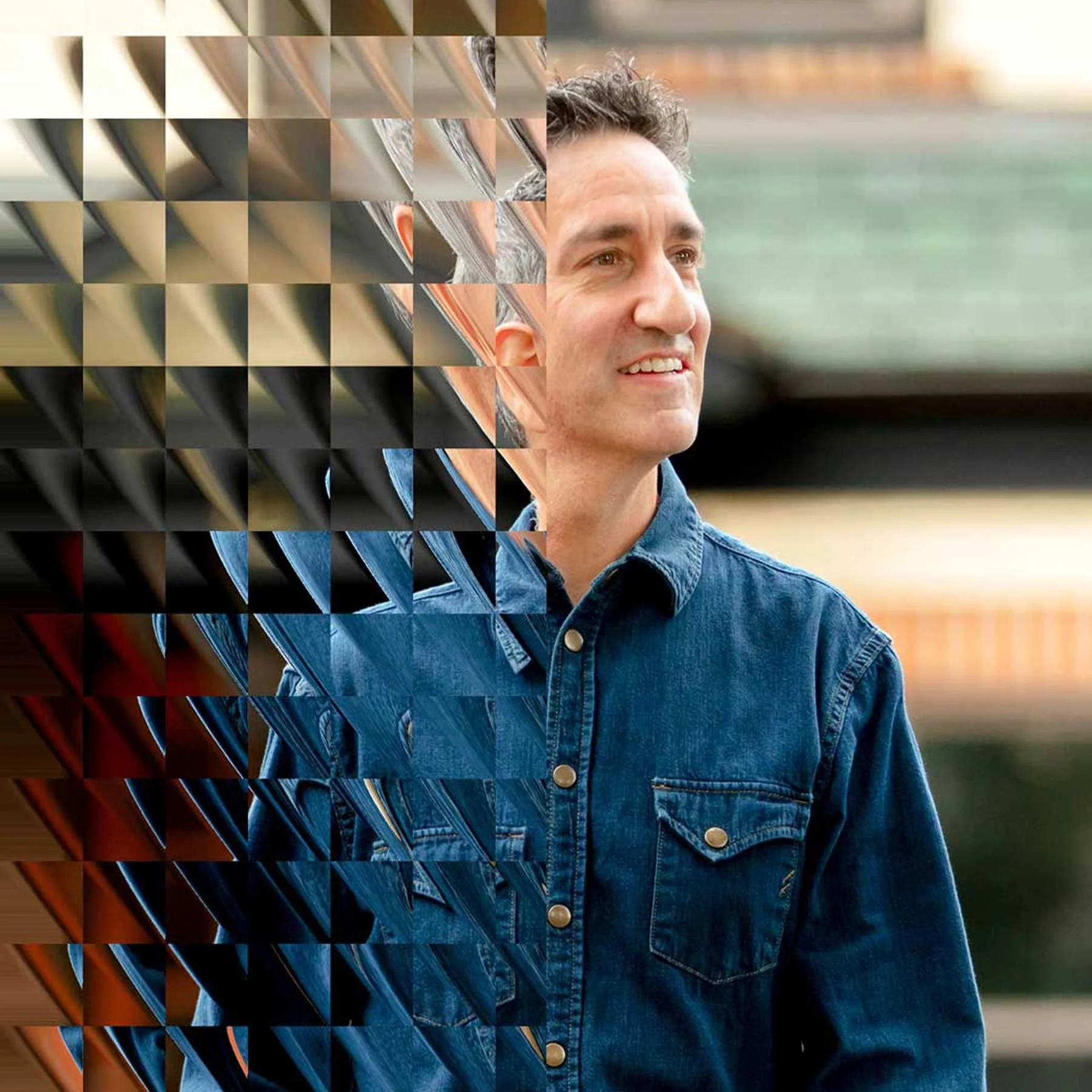 A man wearing a denim jacket stands outdoors, with a background of blurred buildings or structures, smiling and looking off to the side.