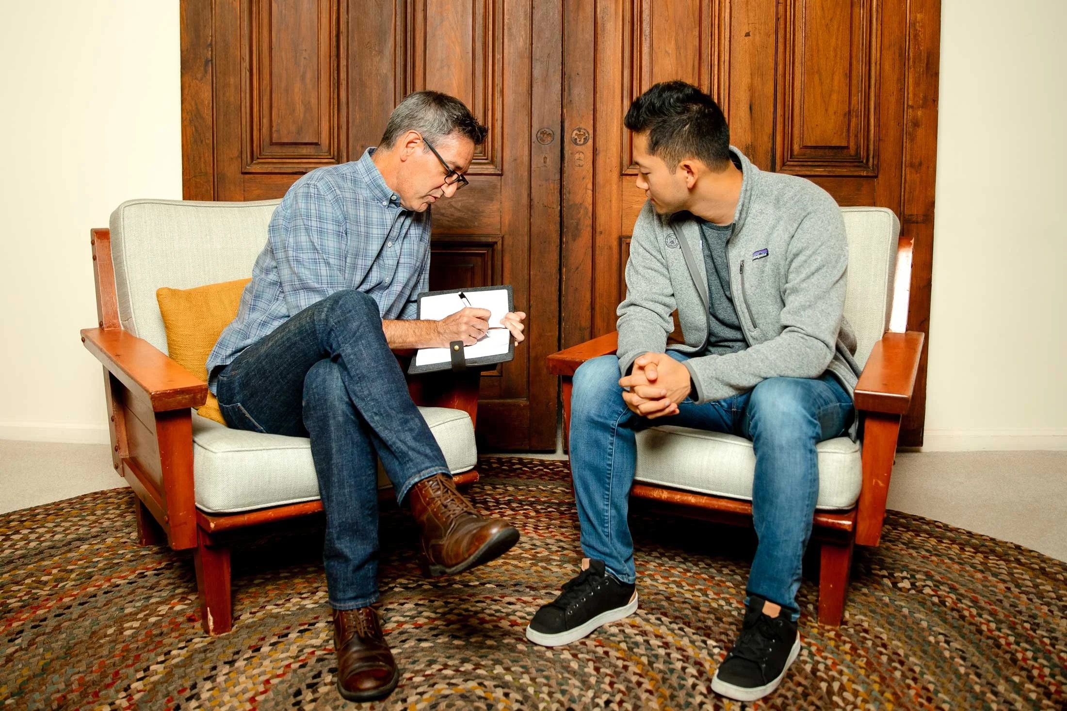 A man in glasses and a plaid shirt taking notes while talking to a young man in a gray jacket and jeans in a therapy session in a living room.