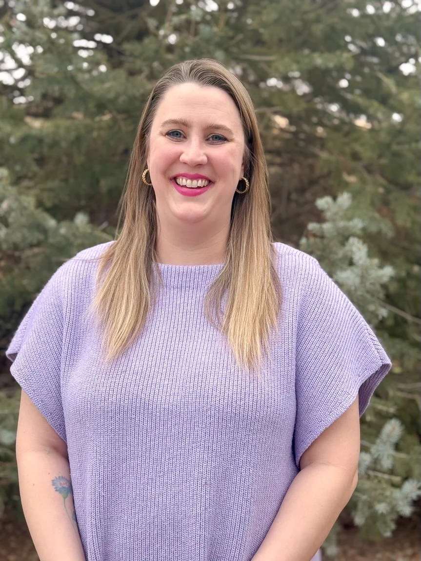 A woman with long blonde hair, wearing a lavender sweater, smiling outdoors with green trees in the background.