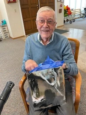 An elderly man smiling while sitting in a wooden chair holding a gift bag.