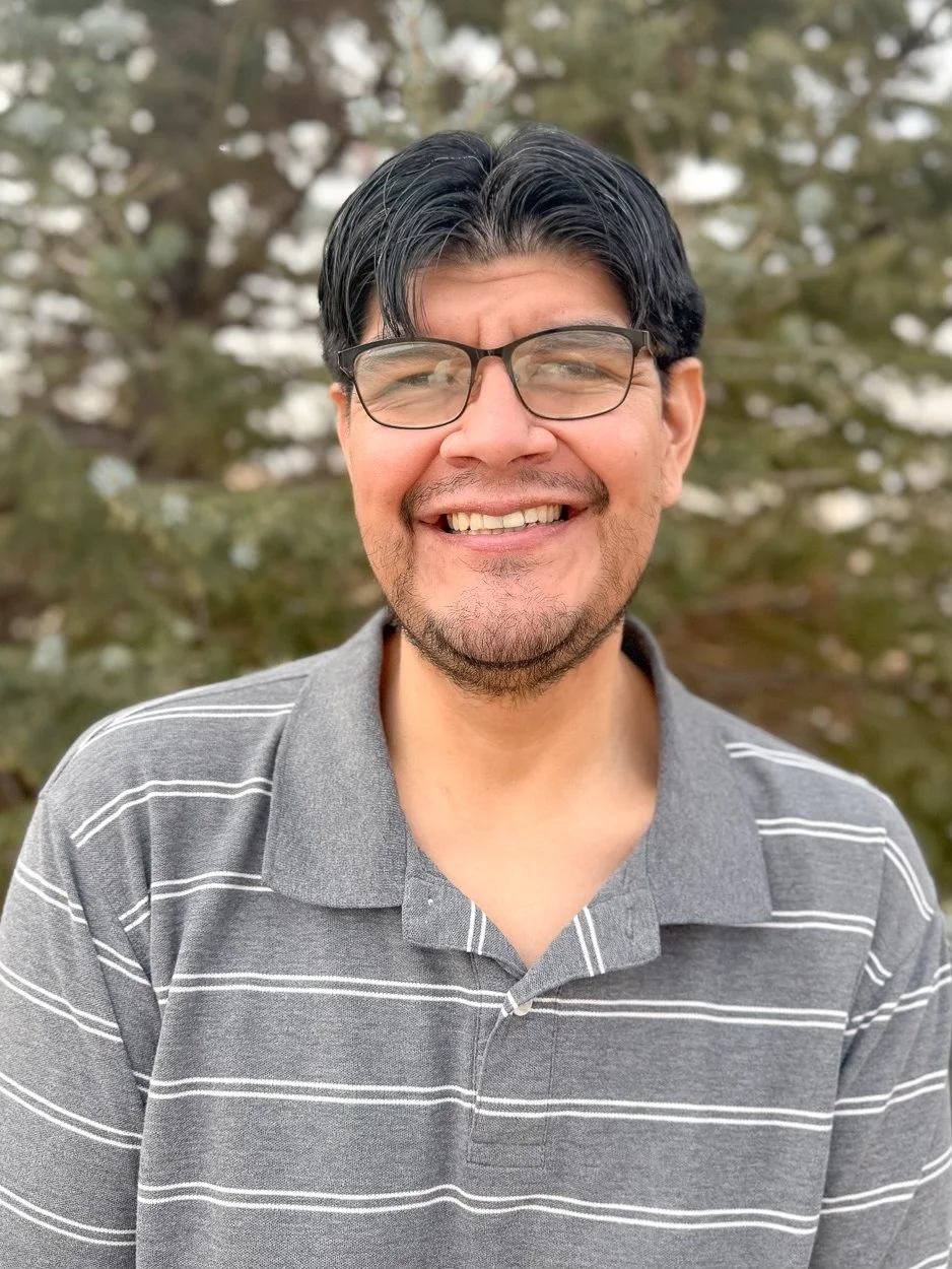 A smiling man with black hair, glasses, and a short beard wearing a gray polo shirt with white horizontal stripes, outdoors in front of blurred trees.