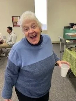 Smiling elderly woman in a blue sweater holding a cup in a communal room.