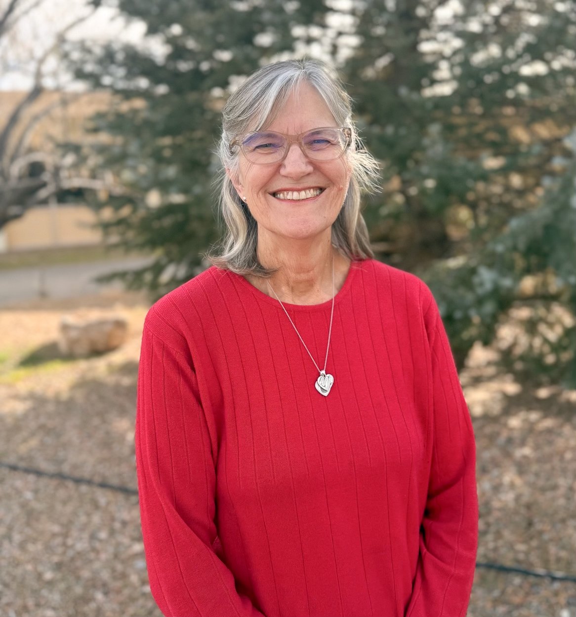An elderly woman with gray hair and glasses, smiling, wearing a red sweater and a heart-shaped necklace, standing outdoors with trees in the background.